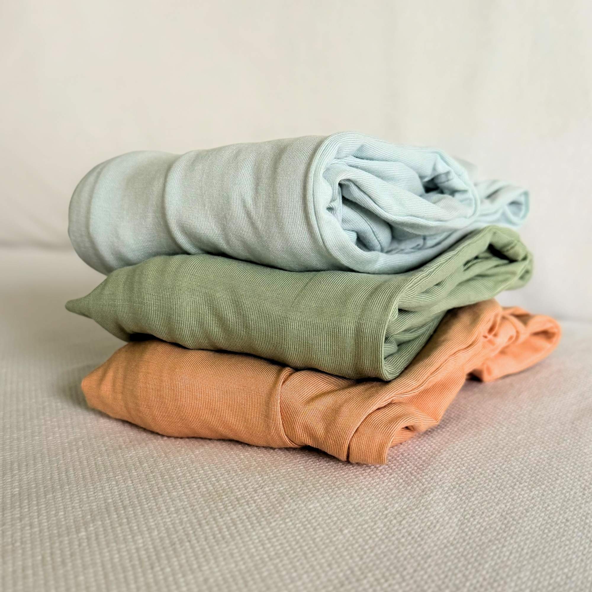 Stack of folded baby grows in light blue, green, and orange on a beige surface.