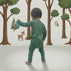 Child in green baby grow playing with a ball in front of a forest wall mural.