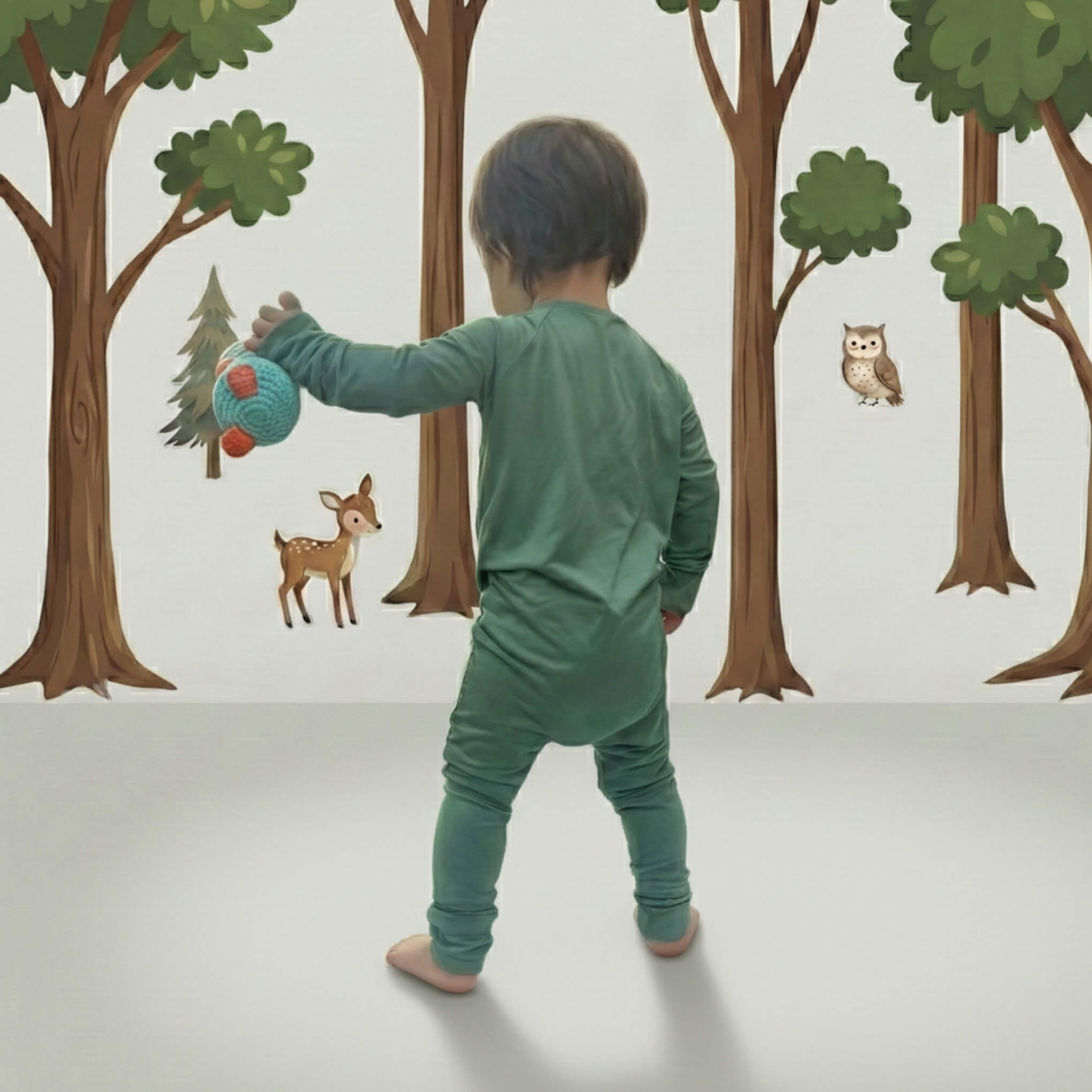 Child in green baby grow playing with a ball in front of a forest wall mural.