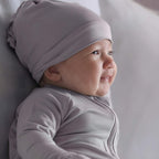 Baby wearing a light gray hat and outfit, lying on a soft surface.